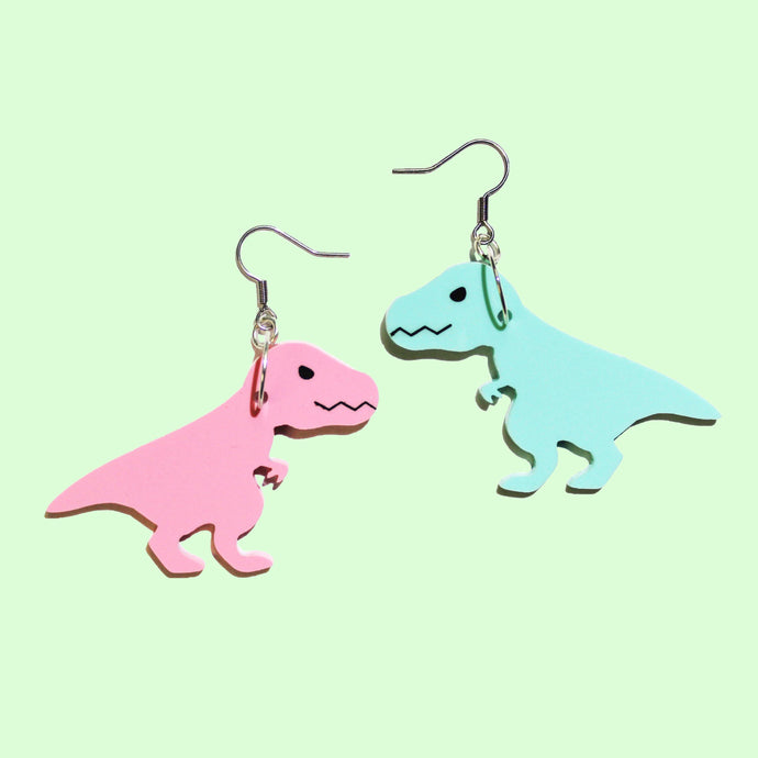 A pair of T Rex earrings. One earring is pink, and the other mint green. Both T Rex are cartoons, with narrowed eyes, jagged teeth and little two fingered hands. The T Rex are a mirror pair. They hang from silver coloured hypoallergenic hooks in front of a pastel pea green background.