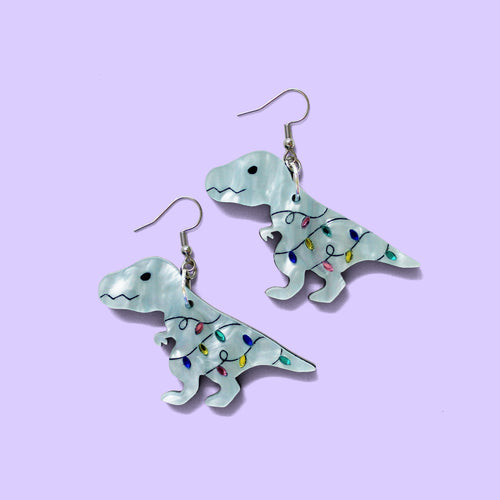 A pair of festive T rex earrings. Oh dear, he's gotten tangled in the Christmas fairy lights. He'd free himself, but you know, the arms are a little too short so... The earrings are made from acrylic and hang from silver-coloured stainless steel hooks. The T rex is mint-coloured with a marble effect. The fairy lights are shiny and reflective in pink, green, yellow and blue.