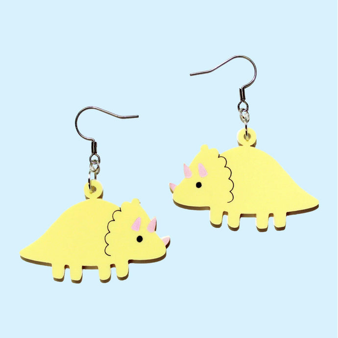 A pair of cute, yellow triceratops earrings. The charms are made from pastel yellow acrylic (plastic) and hang from silver coloured stainless steel hooks. The triceratops are mirror images, each with a scalloped frill and baby pink horns in front of a baby blue background.