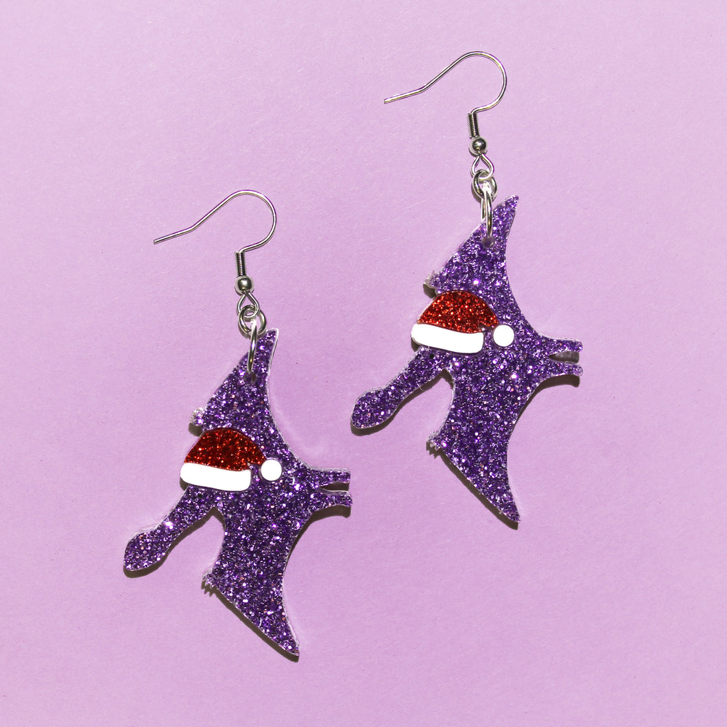 A pair of pterosaur skeleton earrings. Not dinosaur, not pterodactyl, THEY ARE PTEROSAURS. Specifically, Ferrodraco lentoni. The earrings are sparkly purple with sparkly red Santa hats. They have stainless steel hooks which are silver in colour, and they are in front of a purple background.