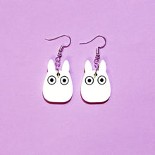 Load image into Gallery viewer, A pair of earrings with silver coloured hooks in front of a purple background. From each hook hangs a white plastic charm that resembles a bunny rabbit with cartoon eyes.
