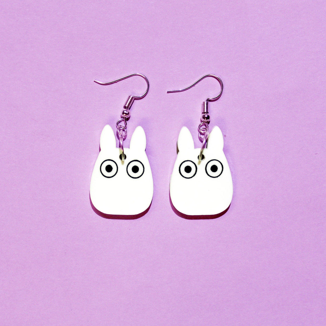 A pair of earrings with silver coloured hooks in front of a purple background. From each hook hangs a white plastic charm that resembles a bunny rabbit with cartoon eyes.