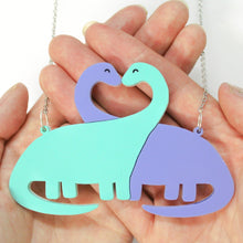 Load image into Gallery viewer, Close up of a dinosaur pendant lying in the palm of two crossed hands. The pendant is of two sauropod dinosaurs with their eyes closed, noses touching and their necks crossed forming sort of a heart shape gap. They are attached on either side to metal rings and a stainless steel chain.

