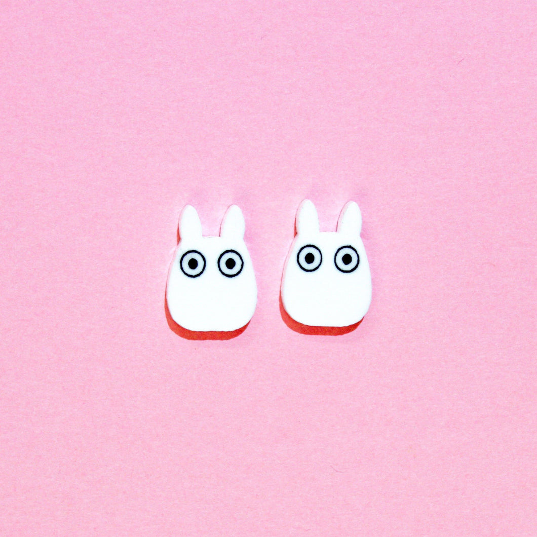 A cute pair of earrings resembling two white rabbits with large cartoon eyes in front of a pink background.