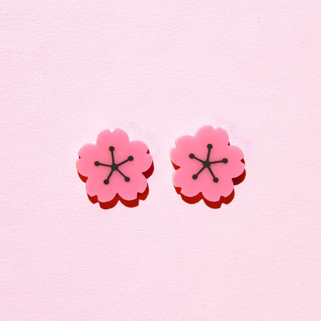 A pair of pink, plastic cherry blossom earrings in front of a pale pink background.