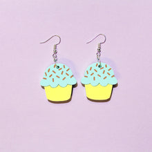 Load image into Gallery viewer, A pair of pastel cupcake earrings on a light purple background with silver coloured hooks. The cupcakes have yellow bottoms and fluffy mint coloured tops with chocolate coloured sprinkles.
