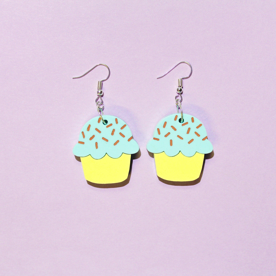 A pair of pastel cupcake earrings on a light purple background with silver coloured hooks. The cupcakes have yellow bottoms and fluffy mint coloured tops with chocolate coloured sprinkles.