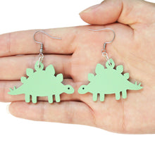 Load image into Gallery viewer, A close up of a hand holding a pair of dinosaur earrings. They are of two green stegosaurus complete with plates and tail spikes. The stegosaurus are mirror images of one another and attached to stainless steel earring hooks.
