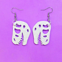 Load image into Gallery viewer, A pair of T-rex skull earrings made from sparkly silvery white glitter acrylic. The Tyrannosaurus skull is a bit cartoonish, with rounded teeth instead of spiky teeth. The earrings are made with stainless steel hooks and are in front of a purple background,

