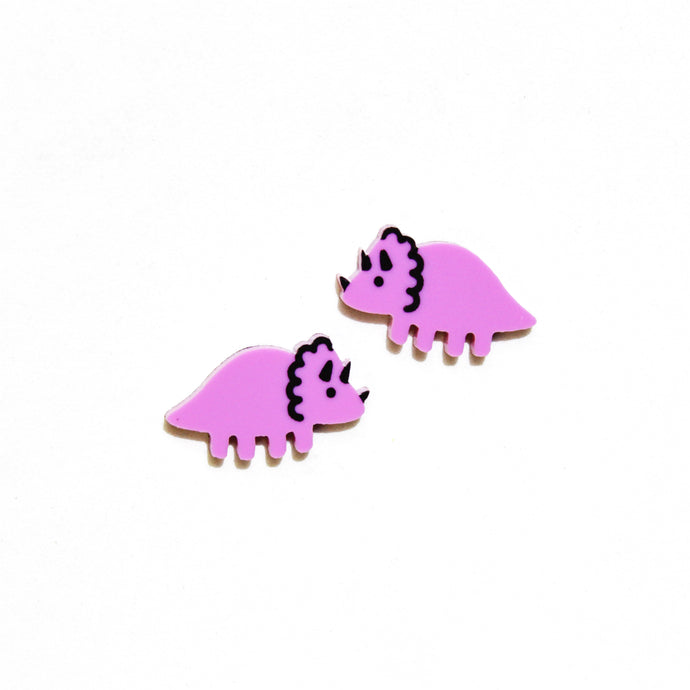 A close up of a cute pair of purple cartoon triceratops stud earrings. The Triceratops or 'Threehorn' dinosaur has two horns near the back of its head, and a little one near the end of its head, and a squiggly frill around its neck. The earrings are small, roughly 2cm long and 1cm tall. The earrings are a pinky purple in front of a white background.