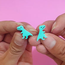 Load image into Gallery viewer, A close up of a pair of mini Tyrannosaurus rex studs being held by two hands. The T rex is cartoonish, in mint green with little stumpy arms and a friendly smile. The hands are in front of a pink background.
