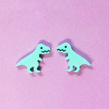 Load image into Gallery viewer, A pair of cartoon T Rex stud earrings. The Tyrannosaurs are mint green with a zigzag mouth, black eyes and little nubs of arms. The earrings are small, about 2cm wide and 1.5 cm tall. The earrings are in front of a lilac purple background.
