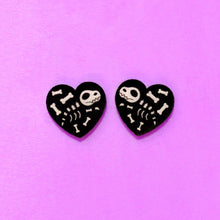 Load image into Gallery viewer, A spoopy pair of heart earrings featuring a dinosaur skeleton! The earrings are black, with slightly transparent white details for the bones and skull. The earrings are small, 1.5 cm tall and 1.5 cm wide and in front of a purple background.
