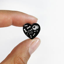 Load image into Gallery viewer, A close up of a heart stud earring featuring a little dinosaur skeleton. The heart is black and the skeleton is a transparent white. The earring is held between the thumb and index finger of a woman in front of a white background.
