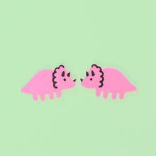 Load image into Gallery viewer, A pair of pink Triceratops stud earrings in front of a pastel green background. The Triceratops have four legs, a muscular tail, three horns on their face and a scalloped frill around their neck. The earrings are small, about 2cm wide and 1cm tall. The Triceratops is pink, with black hand painted details.
