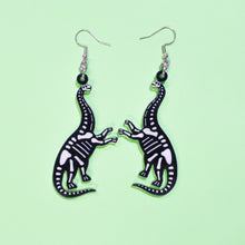 Load image into Gallery viewer, A pair of sauropod skeleton earrings. These earrings are made from acrylic and are black and white, with the silhouette of the animal in black and the bones in a slightly transparent white. The hang from silver coloured stainless steel hooks and are in front of a green background.
