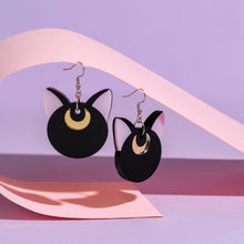 Load image into Gallery viewer, A large pair of black cat head earrings. Minimalist in design, there is no face, just pink ears and shiny gold coloured crescent moons on the forehead. The earrings hang by stainless steel hooks on a pink piece of paper in front of a light purple background.
