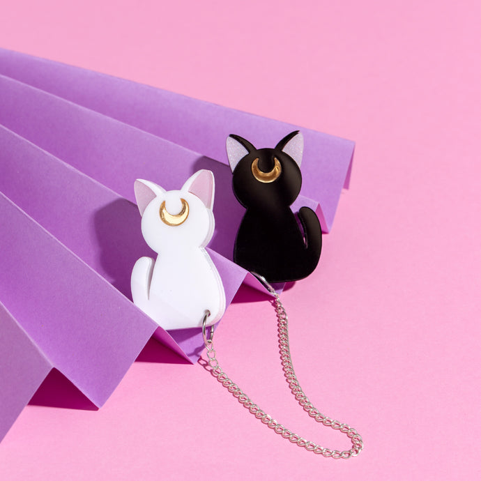 A cardigan clip or two brooches joined together by silver-coloured stainless steel chain. The brooches are a black and white cat, both silhouettes with pink ears and reflective gold coloured crescent moons on their foreheads. The cardigan clip is nestled in a purple, paper fan in front of a pink background.