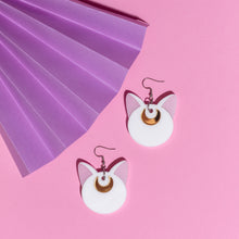 Load image into Gallery viewer, A pair of white cat head earrings with gold coloured crescent moons on their foreheads. The earrings hang from stainless steel hooks in front of a pink and purple background.
