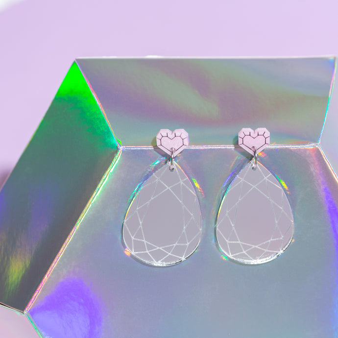A pair of acrylic earrings featuring little pink hearts made to look like cut gemstones at the top and large, pear shaped reflective acrylic with etched detail imitating a gemstone. The earrings sit perched on a holographic background.