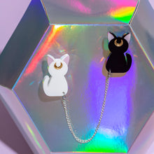 Load image into Gallery viewer, A cardigan clip (two brooches joined together by stainless steel chain). The cardigan clip features two cats, one black, one white, both with pink ears and gold coloured crescent moons on their forehead. The background is a holographic gem made of cardboard.

