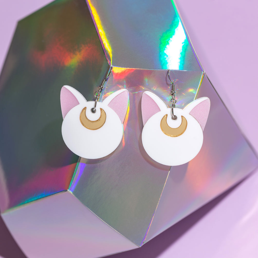 A pair of large cat earrings hanging from a holographic diamond made from paper. The cats are white, with pink ears and gold crescent moons on their foreheads. The earrings hand from silver-tone stainless steel hooks.