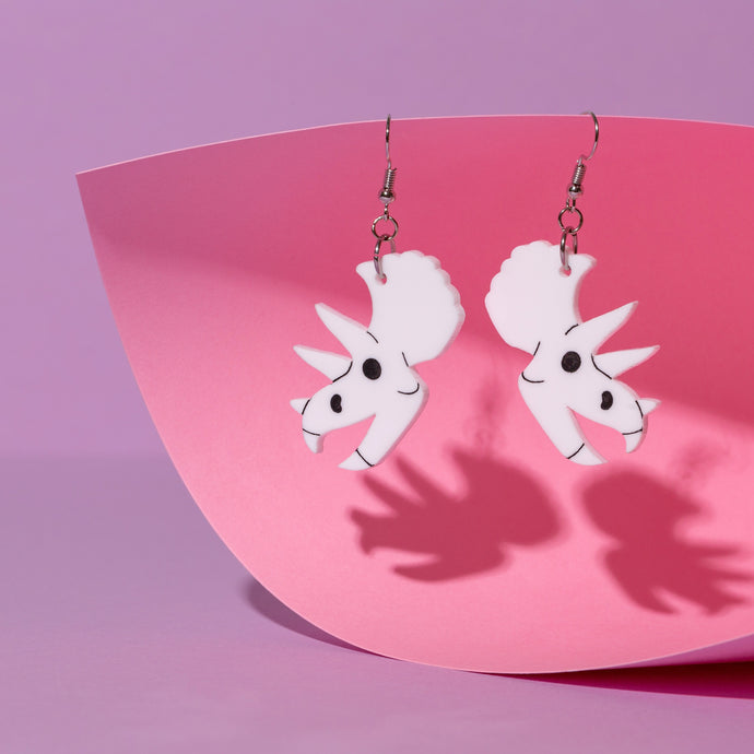 A pair of cartoony Triceratops skull earrings. Not scary at all, this dino has a friendly, goofy expression as well as the frill and three horns you expect to see in this species. The earrings are white with black, hand painted details. They hang from stainless steel hooks. The earrings are in front of a pink and purple background.