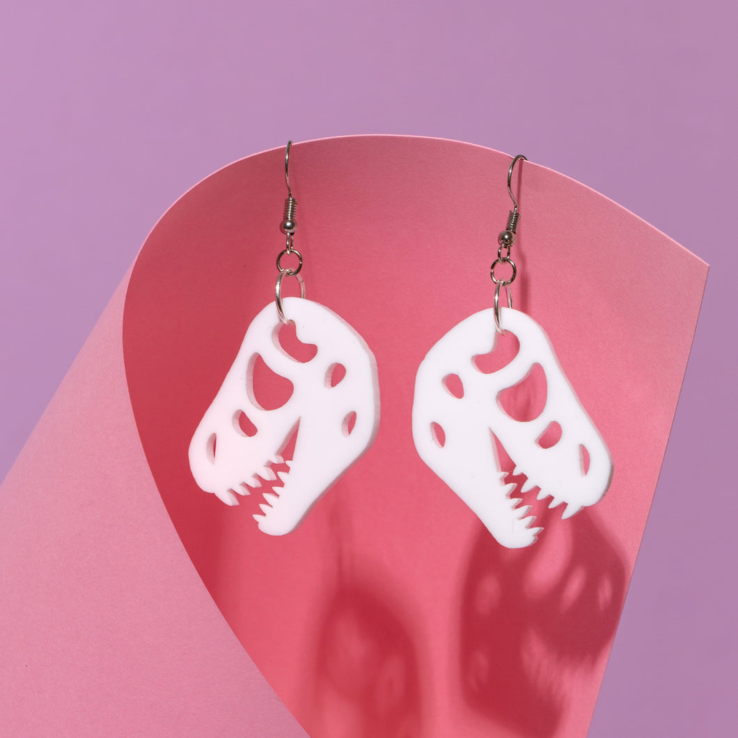 A pair of T Rex skull dangle earrings with stainless steel hooks hanging from a pink piece of card. The earrings are white and are of the skull of Tyrannosaurus rex in white. Behind the pink piece of card is a purple background.