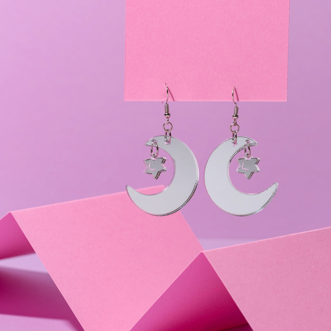 a pair of crescent moon earrings hanging from a pink card. The crescent moons have a little star hanging from them. The moon and star are made from shiny mirror silver acrylic, hanging from silver-coloured stainless steel hooks in front of a purple background.