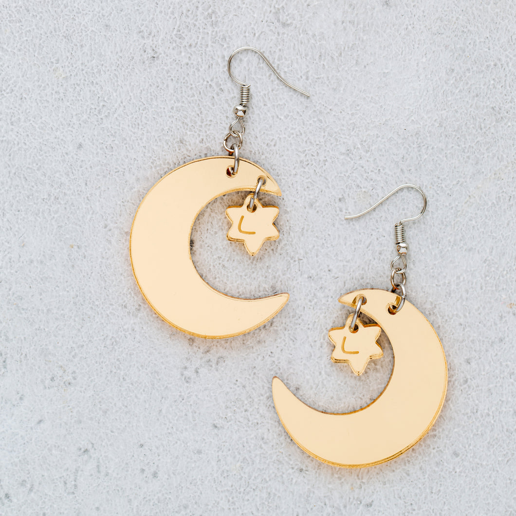 A pair of crescent moon and star earrings on a white tile background. The moon and star are made from reflective gold acrylic and hang from silver-coloured, stainless steel hooks.