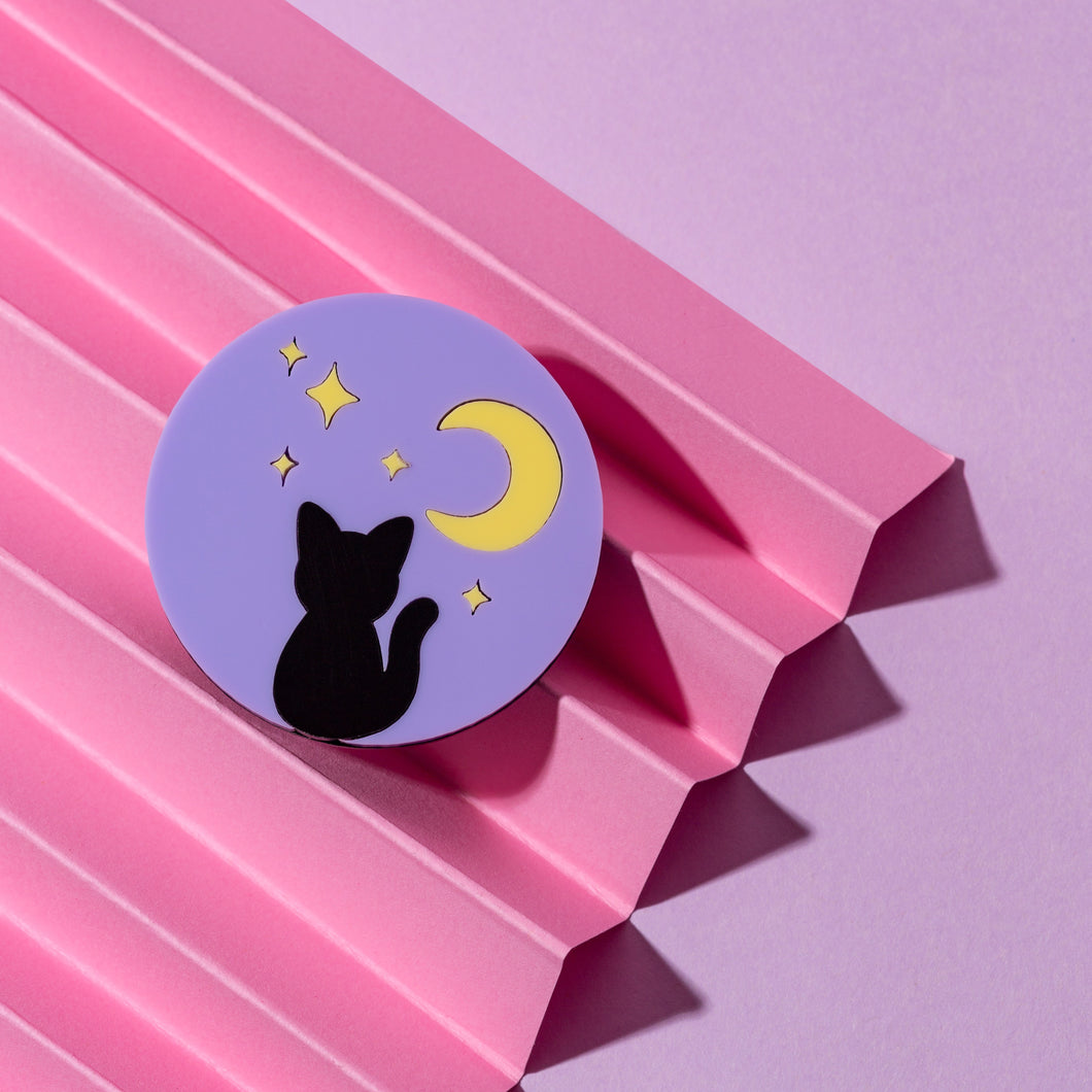 A circle brooch with a purple background, yellow pastel stars and a black cat silhouette. The brooch is 5cm in size and in front of a pink background made from paper.