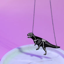 Load image into Gallery viewer, A close up of a plastic dinosaur pendant hanging from a stainless steel chain at either end. The dinosaur is black with white skeleton of an Abelisaurus, a carnivorous dinosaur similar to T rex mid stride with its tail held horizontally. The necklace is in front of a white and purple background.
