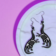 Load image into Gallery viewer, A spooky pair of sauropod skeleton earrings. Sauropods are long necked dinosaurs (think Littlefoot from the Land Before Time movies, Brontosaurus, Brachiosaurus). The earrings are a skeletal reconstruction of a sauropod, so a black silhouette with white transparent bones. The sauropods are rearing up on their hind legs. The earrings are made with acrylic (the dinosaur charm) and stainless steel hooks. The earrings are resting on a white marble plate in front of a purple background.
