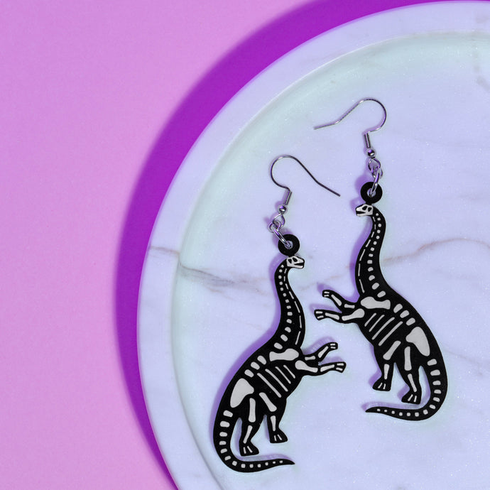 A spooky pair of sauropod skeleton earrings. Sauropods are long necked dinosaurs (think Littlefoot from the Land Before Time movies, Brontosaurus, Brachiosaurus). The earrings are a skeletal reconstruction of a sauropod, so a black silhouette with white transparent bones. The sauropods are rearing up on their hind legs. The earrings are made with acrylic (the dinosaur charm) and stainless steel hooks. The earrings are resting on a white marble plate in front of a purple background.