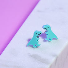 Load image into Gallery viewer, A pair of stud earrings based on the Late Cretaceous dinosaur, Tyrannosaurus rex. The T rex is spearmint green, with hand painted eyes and mouth. The mouth is a zig zag shape and looks a bit like it&#39;s smiling. The stainless steel posts and backs can be seen behind the T-rex. The earrings are in front of a sparkly white and purple background.
