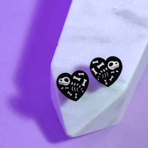 A spooky pair of heart earrings with a little dinosaur skeleton in transparent white. The earrings are small, 1.5cm by 1.5cm and balanced on a sparkly white crystal on a purple background. The earrings are black with white transparent bones including a cartoonish skull.