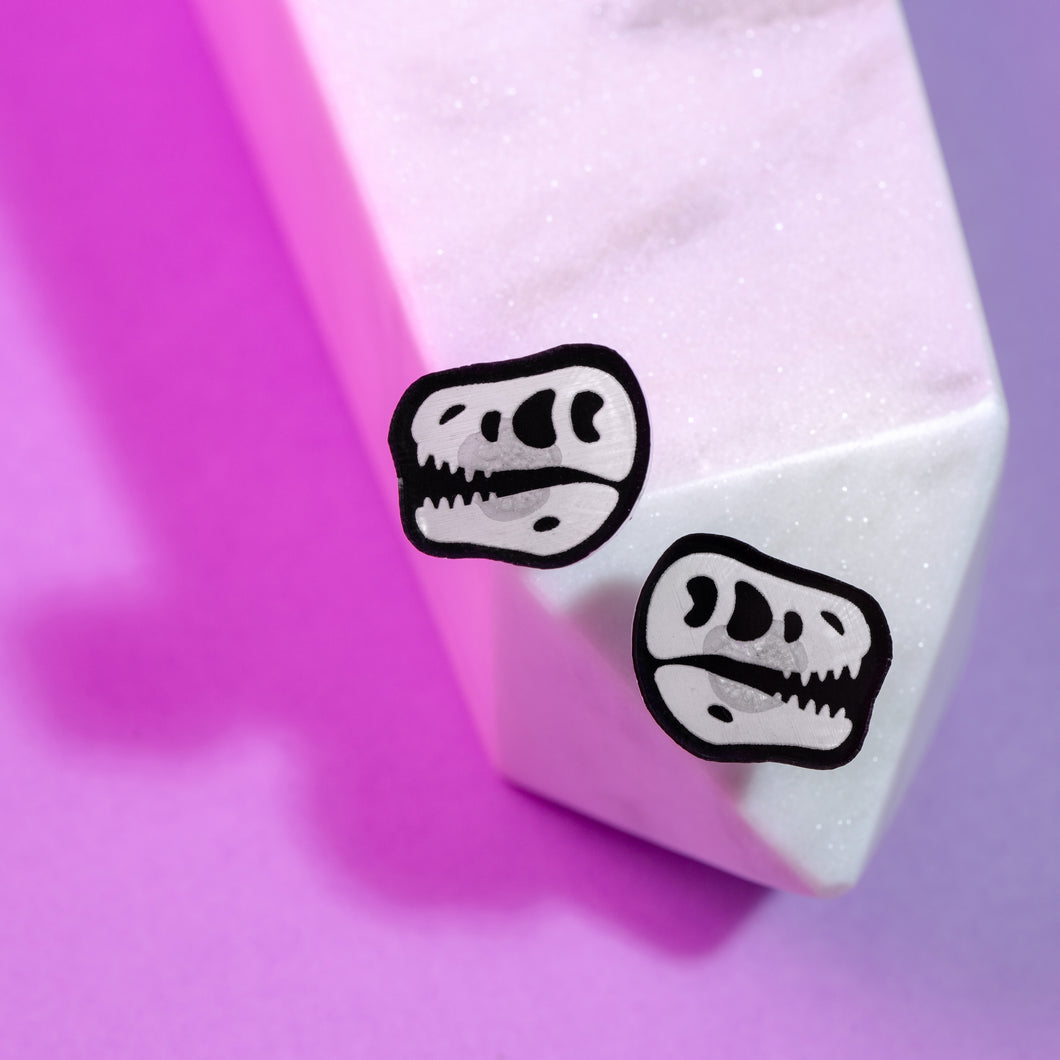 A pair of cute and spooky T rex skull stud earrings. The earrings are made from acrylic, which is a type of plastic, and are black and white (transparent white with black details and black silhouette). There are roughly circular marks behind the skull design. The earrings are small and balanced on the end of a shimmery white crystal in front of a purple background.