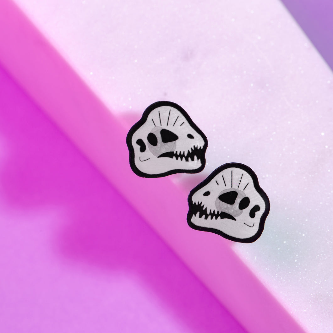 Acrylic Earrings based on the skull of the Early Jurassic dinosaur, Dilophosaurus. The skull is in a transparent white, with black detail. There is a circular shadow behind the skull in the centre of the skull design. The earrings are small, with a glittery white and purple-pink background.