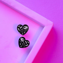 Load image into Gallery viewer, A pair of black heart studs, with transparent white cartoon dinosaur skeletons and bones. The earrings are small, about 15mm by 15mm in size, and made from acrylic, which is a type of plastic. The earrings are in front of a pink and purple background.
