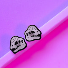 Load image into Gallery viewer, Dinosaur skull earrings of the Early Jurassic carnivore from North America, Dilophosaurus! Dilophosaurus is best known from Jurassic Park and was depicted in that movie with a frill and spitting venom. The earrings are black and transparent white, and in front of a purple and pink background.
