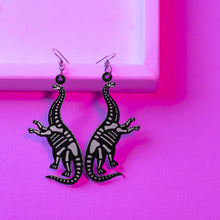 Load image into Gallery viewer, A spooky pair of dinosaur skeleton earrings. The dinosaurs are sauropods, which are herbivores and the largest land animals to walk on the surface of the Earth. The design has them posed standing on their back legs, with their front legs raised. The earrings are black, with transparent white bones detailing. The earrings are made with hypoallergenic stainless steel hooks and are in front of a pinky purple background.
