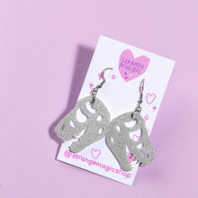 Load image into Gallery viewer, A pair of glittery dinosaur skull earrings. The dinosaur skull is based on T-rex, or Tyrannosaurus rex and hangs from silvertone stainless steel hooks mounted onto a card that says Strange Magic in a pink heart, and @strangemagicshop on the bottom. The card is in front of a light purple background.
