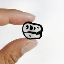 Load image into Gallery viewer, A close up of a T rex stud earring. The earring is of a Tyrannosaurus skull in black and white, with the bones in a transparent white. The shadow of the post can be seen a little bit through the skull. The earring is small and held in the fingers of a woman in front of a white background.
